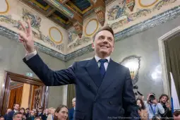 Mężczyzna w garniturze z uśmiechem, wykonujący gest zwycięstwa w eleganckim wnętrzu.