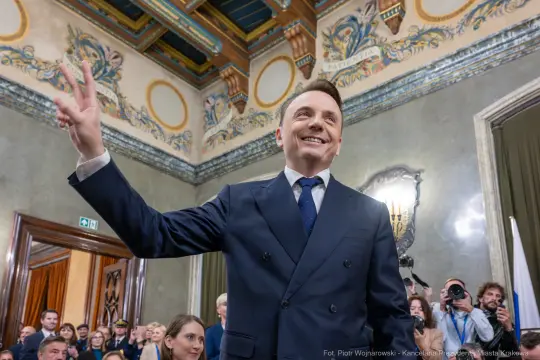 Mężczyzna w garniturze z uśmiechem, wykonujący gest zwycięstwa w eleganckim wnętrzu.