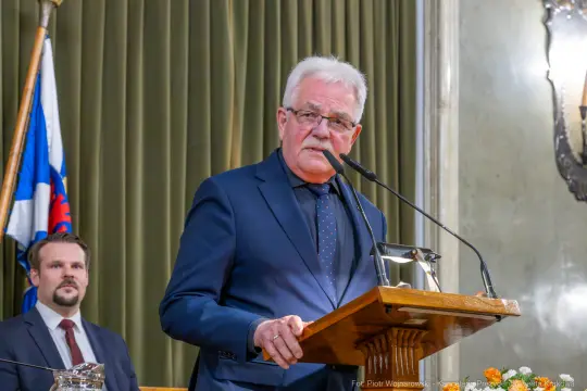 Mężczyzna przemawia z mównicy, w tle flagi i inny mężczyzna.