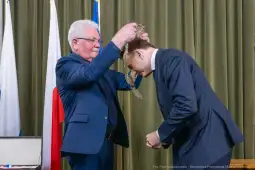 Wręczenie odznaczenia przez mężczyznę w okularach drugiemu mężczyźnie w garniturze.