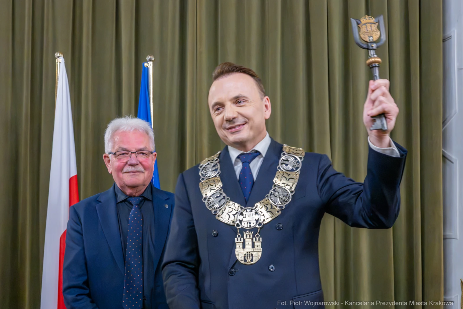 Mężczyzna w garniturze z łańcuchem prezydenckim trzyma berło, obok stoi inny mężczyzna.  Autor: Piotr Wojnarowski