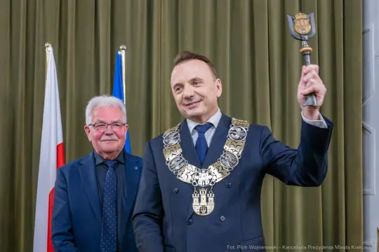 Mężczyzna w garniturze z łańcuchem prezydenckim trzyma berło, obok stoi inny mężczyzna.