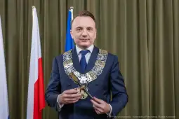 Mężczyzna w garniturze z łańcuchem prezydenckim, stojący przed flagami Polski i Krakowa.