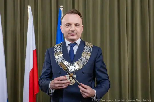 Mężczyzna w garniturze z łańcuchem prezydenckim, stojący przed flagami Polski i Krakowa.