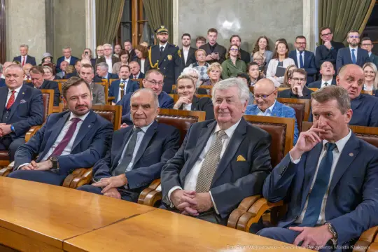 Grupa mężczyzn w garniturach siedzących w sali obrad, w tle publiczność.