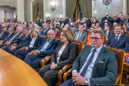 Grupa ludzi siedzących w sali, w tle widoczni goście i uczestnicy wydarzenia.