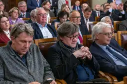 Grupa ludzi siedzących w sali, niektórzy z nich wpatrzeni w swoje telefony.