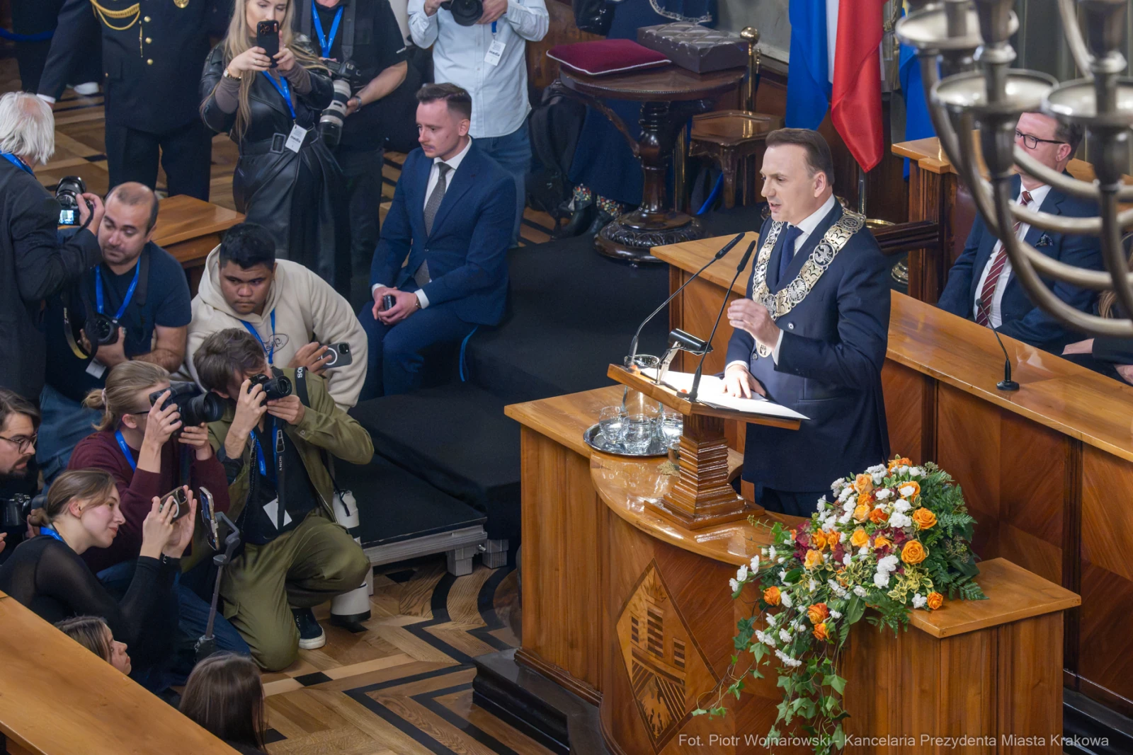 Mężczyzna w garniturze przemawia z mównicy, otoczony dziennikarzami i flagami.  Autor: Piotr Wojnarowski