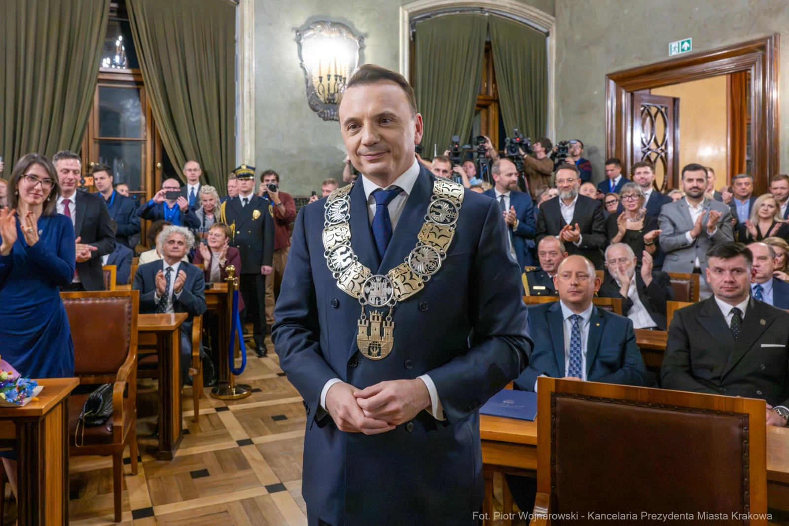 Mężczyzna w eleganckim garniturze z łańcuchem na szyi, otoczony przez zgromadzonych gości.  Autor: Piotr Wojnarowski