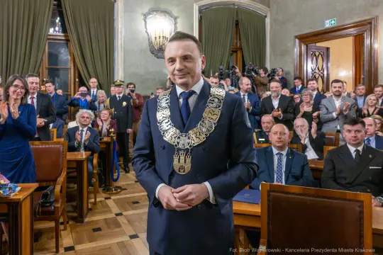 Mężczyzna w eleganckim garniturze z łańcuchem na szyi, otoczony przez zgromadzonych gości.