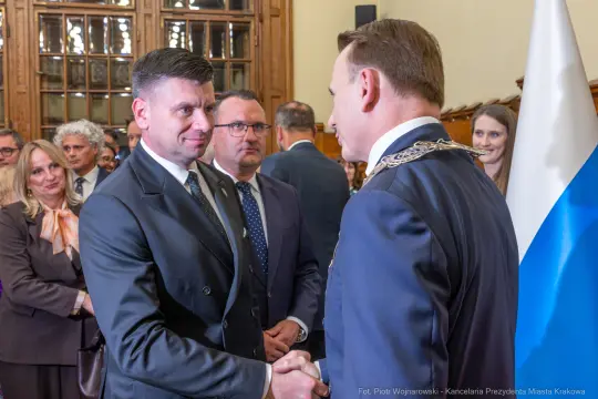 Spotkanie dwóch mężczyzn w garniturach, uściski dłoni, w tle goście i flagi.
