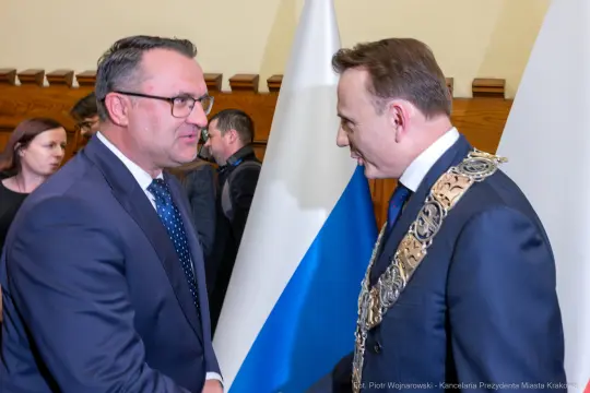 Dwaj mężczyźni w garniturach rozmawiają, w tle flagi i osoby w formalnych strojach.