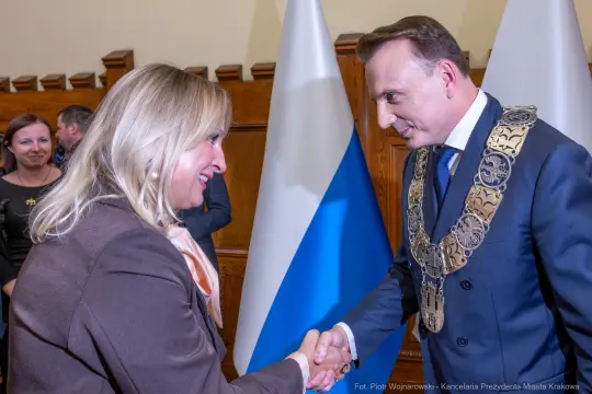 Spotkanie dwóch osób w formalnej atmosferze, uścisk dłoni. W tle flagi.