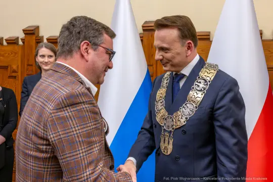 Spotkanie dwóch mężczyzn w garniturach, jeden z łańcuchem prezydenckim, w tle flagi.