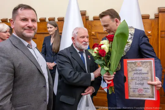 Mężczyzna z kwiatami i dyplomem, wręczający podziękowania innemu mężczyźnie. W tle inni uczestnicy wydarzenia.