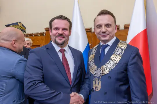Dwaj mężczyźni w garniturach, uściski dłoni, flagi Polski i Rosji w tle.