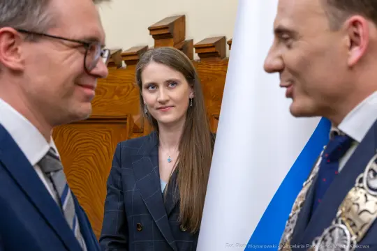 Trzech ludzi w formalnych strojach, rozmowa, kobieta w tle z zainteresowaniem obserwuje.