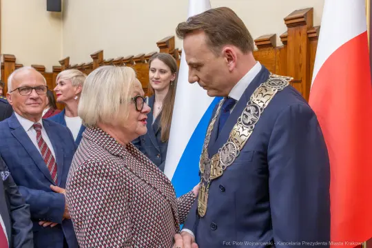 Spotkanie dwóch osób w formalnych strojach, w tle flagi i inni uczestnicy wydarzenia.