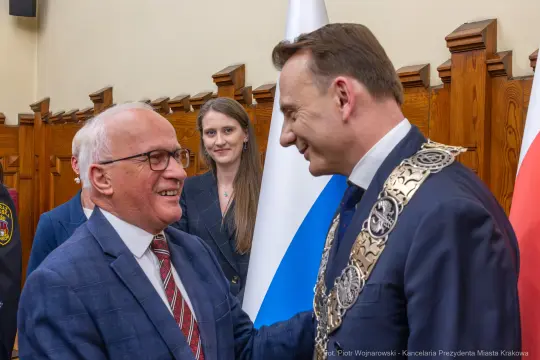 Spotkanie dwóch mężczyzn w garniturach, z uśmiechami, w tle flagi Polski i Krakowa.