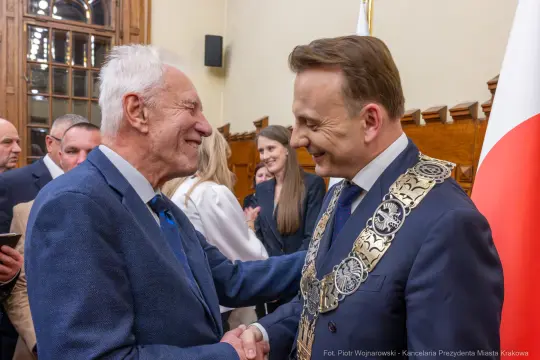 Dwaj mężczyźni uścisnęli sobie dłonie, w tle flagi i osoby w formalnych strojach.