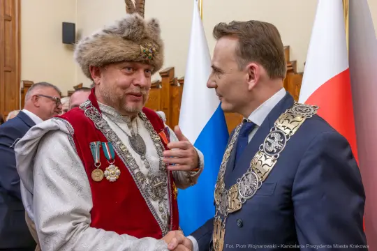 Dwaj mężczyźni w eleganckich strojach rozmawiają, jeden z nich w tradycyjnym nakryciu głowy.