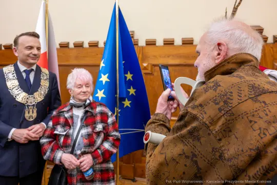 Mężczyzna robi zdjęcie starszej kobiecie i mężczyźnie w oficjalnych strojach, w tle flagi Polski i UE.