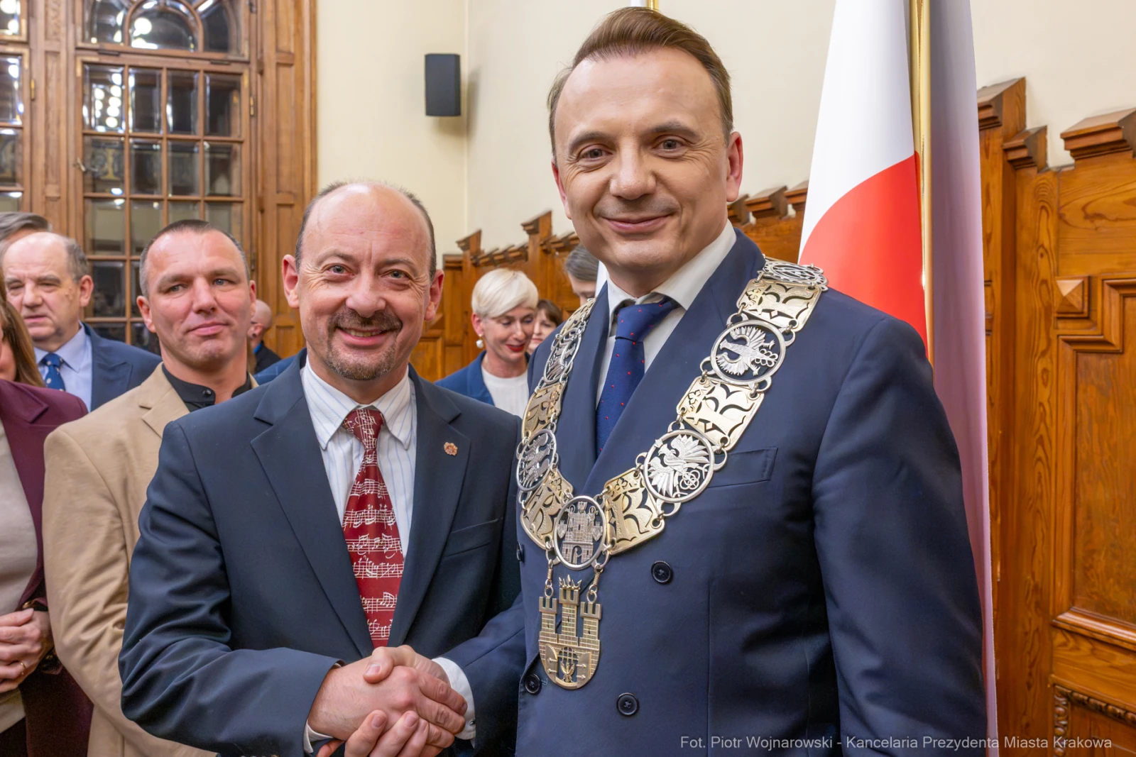 Dwóch mężczyzn uścisnęło sobie dłonie w eleganckim wnętrzu z flagą Polski w tle.  Autor: Piotr Wojnarowski