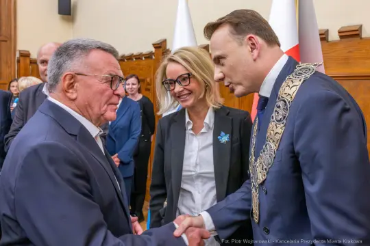 Spotkanie dwóch mężczyzn w garniturach, uściski dłoni, kobieta w tle.