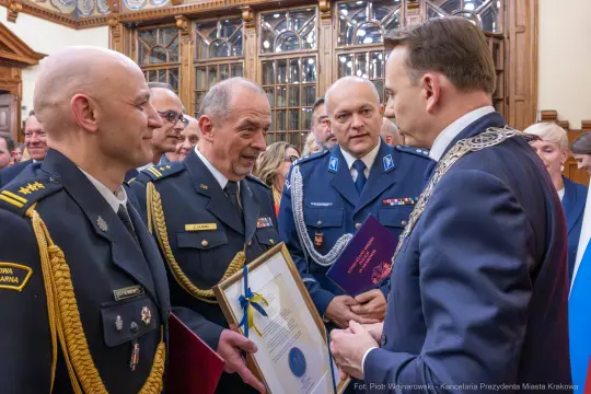Wręczenie odznaczeń w sali z dekoracyjnymi oknami, mężczyźni w mundurach rozmawiają.