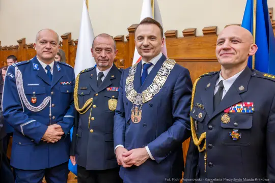 Czterech mężczyzn w mundurach, jeden z łańcuchem honorowym, w sali z flagami.