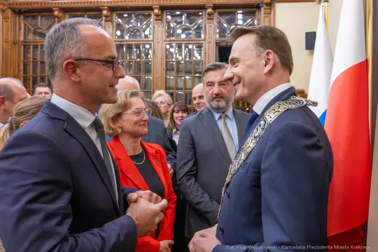 Spotkanie dwóch mężczyzn w formalnych strojach, w tle flagi i goście w eleganckich ubraniach.