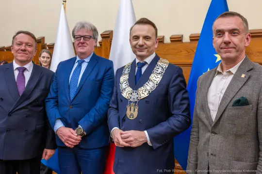 Czterech mężczyzn w garniturach, jeden z łańcuchem prezydenckim, w sali z flagami.