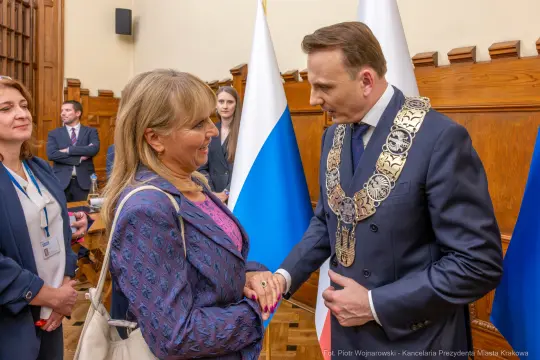 Spotkanie dwóch osób w formalnym otoczeniu, uściski dłoni i flagi w tle.