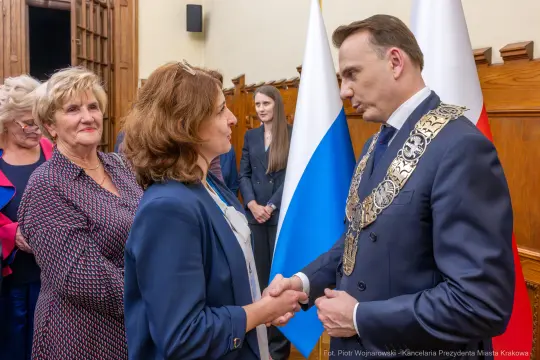 Spotkanie dwóch osób z uściskiem dłoni, w tle flagi i uczestnicy wydarzenia.