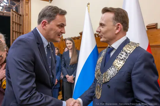 Dwaj mężczyźni w garniturach uścisną ręce, w tle widoczni inni uczestnicy spotkania.