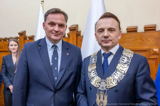 Dwaj mężczyźni w garniturach, jeden z łańcuchem, w tle flagi i drewniane panele.