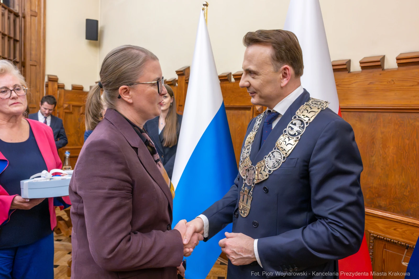 Uścisk dłoni między dwiema osobami, w tle flagi i osoby w formalnych strojach.  Autor: Piotr Wojnarowski