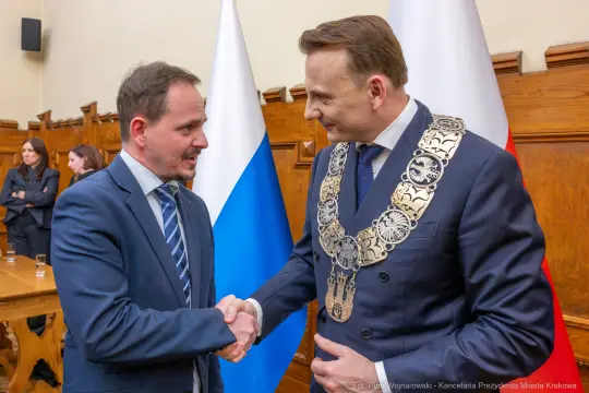 Dwaj mężczyźni w garniturach, jeden z łańcuchem prezydenckim, uściski dłoni. Flagi w tle.