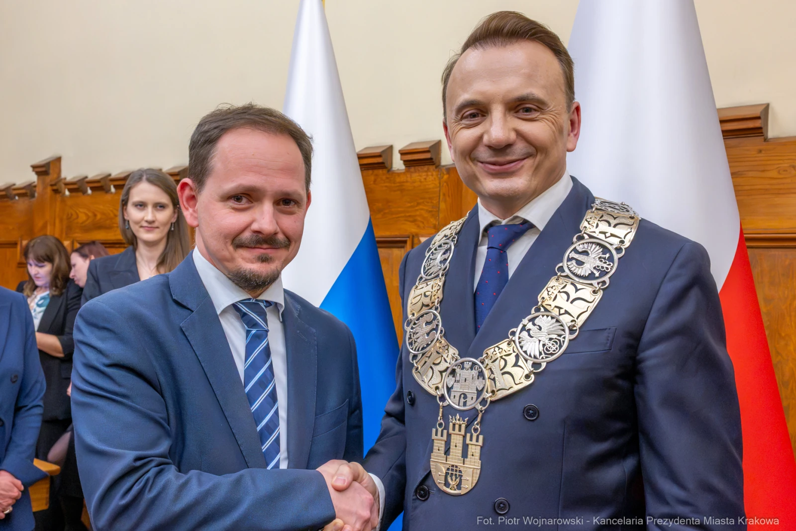 Dwaj mężczyźni uścisnęli sobie dłonie, jeden z nich w łańcuchu prezydenckim. W tle kobiety i flagi.  Autor: Piotr Wojnarowski
