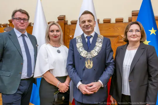 Cztery osoby w formalnych strojach, w tym mężczyzna z łańcuchem prezydenckim, w sali z flagami.