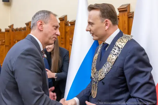 Dwaj mężczyźni w garniturach wymieniają uścisk dłoni, w tle flagi i elementy dekoracyjne.