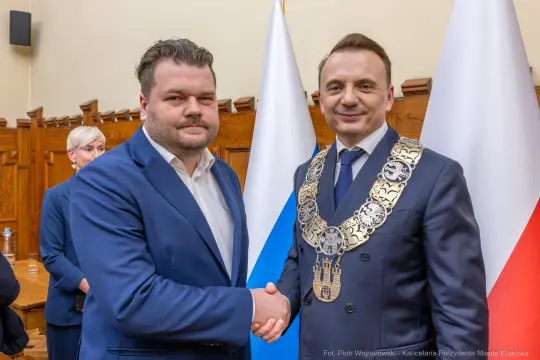 Dwóch mężczyzn w garniturach uścisnęło sobie dłonie, w tle flagi Polski i Krakowa.