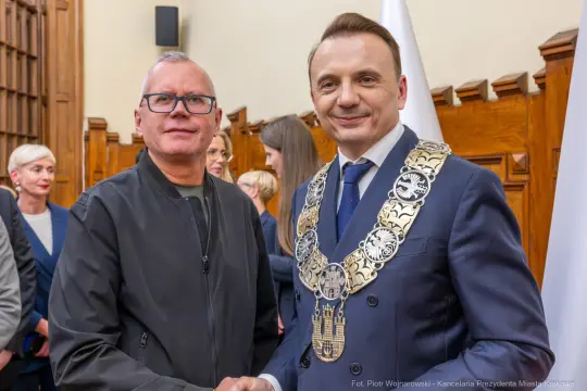 Mężczyźni w garniturach uścisną ręce, w tle flagi i osoby w formalnych strojach.