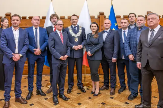 Grupa ludzi w formalnych strojach, w tym burmistrz w łańcuchu, na tle flagi Polski i Unii Europejskiej.