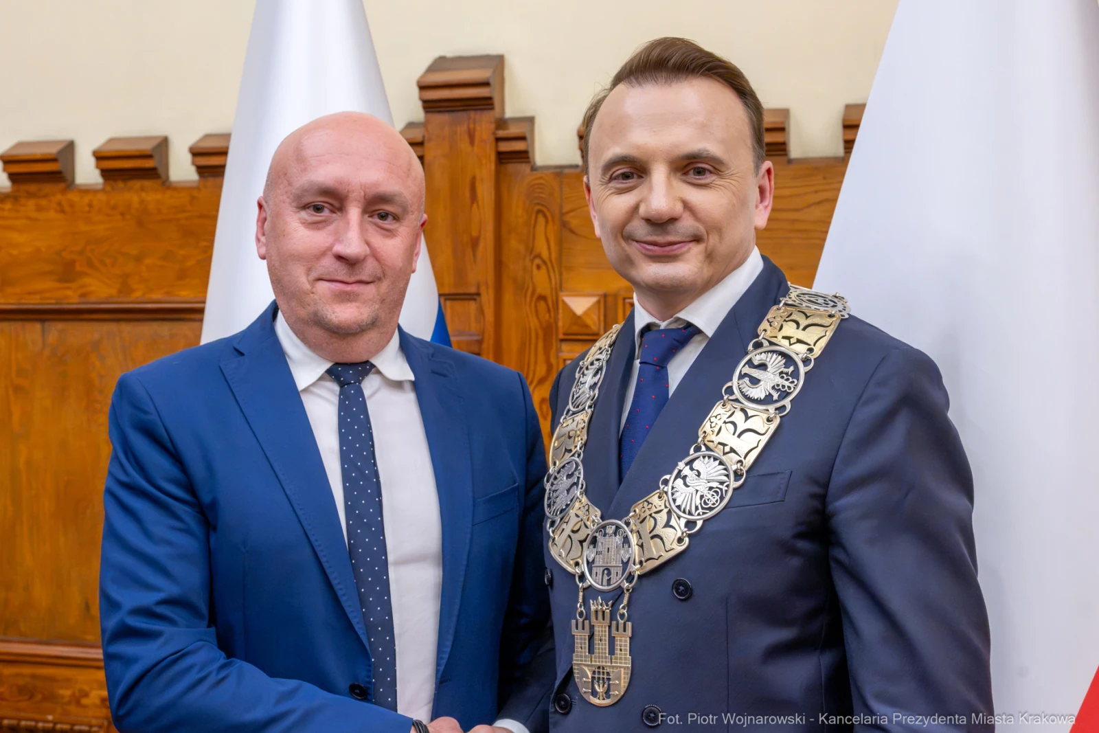 Dwóch mężczyzn w garniturach, uściski dłoni, symboliczne łańcuchy na szyi, flagi w tle.  Autor: Piotr Wojnarowski