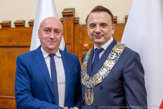 Dwóch mężczyzn w garniturach, uściski dłoni, symboliczne łańcuchy na szyi, flagi w tle.