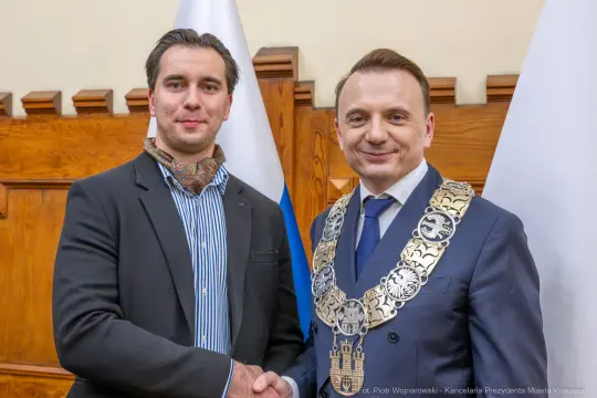 Dwaj mężczyźni w garniturach, jeden z łańcuchem prezydenckim, uściski dłoni. Flagi w tle.