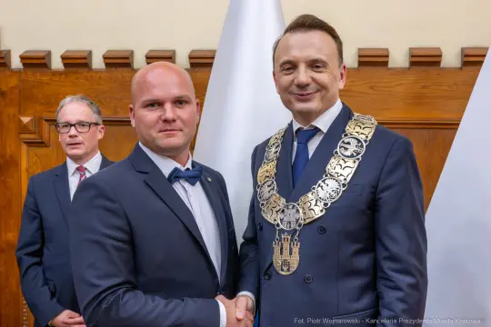 Mężczyzna w garniturze z odznaczeniem uścisnął dłoń drugiemu mężczyźnie w garniturze. W tle widoczny trzeci m