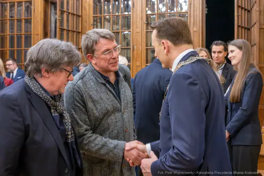 Spotkanie dwóch mężczyzn w garniturach, uścisk dłoni, w tle inni uczestnicy wydarzenia.