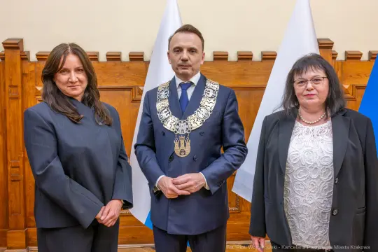 Trzy osoby w formalnych strojach, w tle flagi i drewniana ściana.